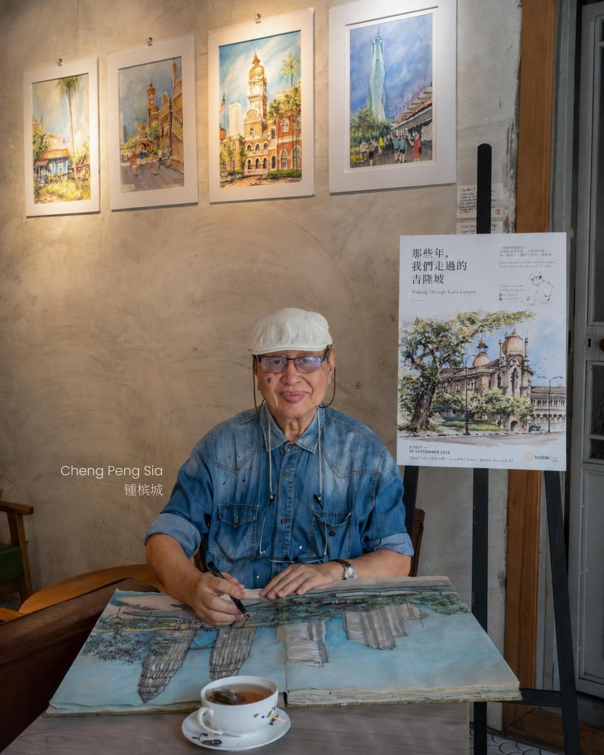 The artist sketching at a café table, a cup of coffee beside him and framed watercolours on the wall behind